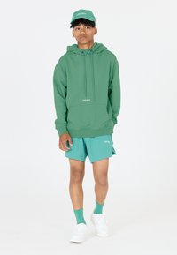 Grüne Kapuzenjacke mit Kordelzügen, kombiniert mit türkisfarbenen Shorts, passenden Socken und weißen Sneakers. Logo "LES ARCS" auf beiden.