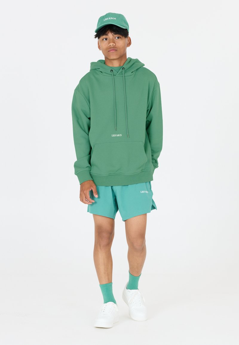 Grüne Kapuzenjacke mit Kordelzügen, kombiniert mit türkisfarbenen Shorts, passenden Socken und weißen Sneakers. Logo "LES ARCS" auf beiden.