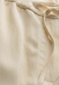 Nahaufnahme von beigem Stoff mit sichtbarer Webstruktur und einer genähten Naht neben einer Stoffschlaufe oder einem Stoffband.