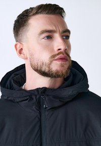 Chaqueta con capucha negra, con cremallera en la parte delantera, fabricada en un tejido suave y con un cordón ajustable en la capucha. El modelo muestra sus rasgos faciales sin accesorios.