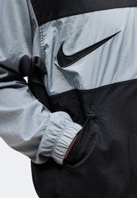 Άτομο που φοράει μαύρο και γκρι αδιάβροχο Nike με το χέρι στην μπροστινή τσέπη, δείχνοντας το ελαστικό μανίκι και το λογότυπο Nike στο στήθος.