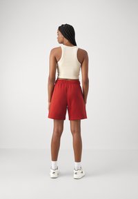 Shorts en coton rouges avec une taille élastique associés à un débardeur crème. Le mannequin se tient sur un fond neutre, portant des chaussettes blanches et des baskets.