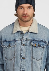Homme portant un bonnet en tricot bleu et une veste en denim bleu clair avec un col en sherpa blanc, faisant face à l'avant sur un fond uni.