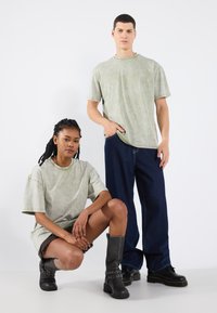 Pár modelů oblečených v oversized světle zelených tričkách, černých džínách a černých botách; předvádějících uvolněný střih a odřenou texturu.