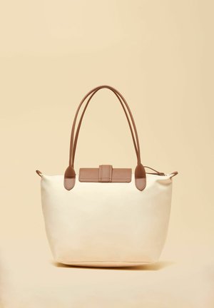 Kreemikas kandekott pruuni nahast käepidemete ja klapiga, neutraalse beežika taustaga.