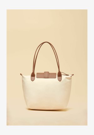 Kreemikas kandekott pruuni nahast käepidemete ja klapiga, neutraalse beežika taustaga.