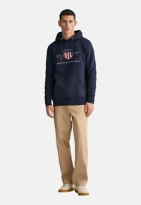 Felpa blu navy con logo frontale, cordini e tasca a marsupio, abbinata a pantaloni beige e sneakers bianche con suola in gomma.