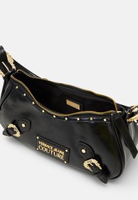 Czarna torebka ze skóry lakierowanej z złotymi okuciami, zamkiem błyskawicznym, ozdobnymi ćwiekami i wyraźną tabliczką z logo. Kompaktowy, zakrzywiony kształt.