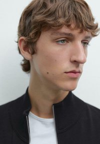 Profilansicht eines jungen Mannes mit hellbraunem, welligem Haar, der eine schwarze Zip-Jacke über einem weißen Hemd trägt. Die Haut hat eine glatte Textur und minimalen Gesichtshaarwuchs.