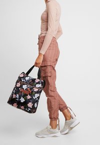 Henkilö, jolla on päällään vaaleanpunainen pitkähiha, raitahousut, kantaa mustaa kukallista Puma-kangaskassia ja beigen värisiä Puma-slipsneakers-kenkiä.