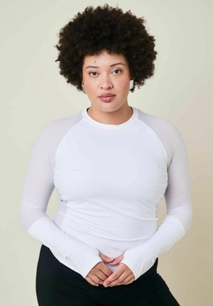 Femme aux cheveux bouclés portant un haut en maille blanc à manches longues et un pantalon noir, debout, mains jointes devant, fond neutre.
