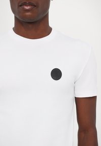 Hvid bomuld t-shirt med et rundt sort lædermærke med prægede logo på venstre bryst. Korte ærmer med klassisk rund hals.