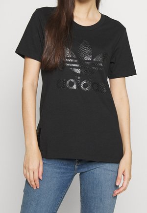 Woman wearing black Adidas t-shirt with shiny trefoil logo and blue jeans, standing against a plain light background.