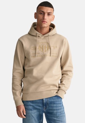 Beige hoodie met trekkoorden en een logo aan de voorkant, voorzien van een kangoeroezak. Draagt in combinatie met lichtblauwe jeans; de stof lijkt zacht en gestructureerd.