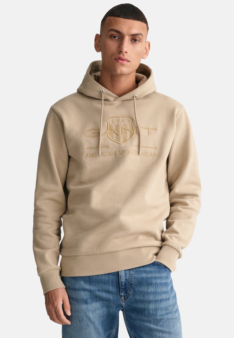 Felpa beige con coulisse e logo frontale, dotata di tasca a marsupio. Indossata con jeans azzurri chiari; il tessuto appare morbido e testurizzato.