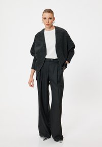 Cardigan oversize grigio scuro indossato sopra una camicia bianca, abbinato a pantaloni neri a vita alta. Scarpe argentate a punta notevoli.