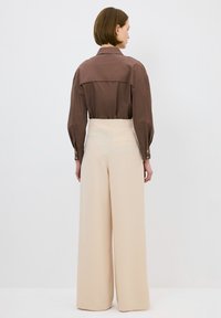 Camicia marrone con colletto e maniche lunghe; pantaloni beige a vita alta e a gamba larga con tasca posteriore. Tessuto liscio e leggero.