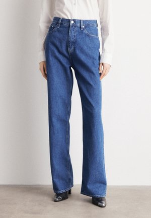 Femme portant un jean bleu taille haute à jambes droites, une chemise blanche à manches longues et des bottes noires à bout pointu, debout sur un sol gris.