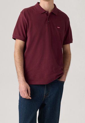 Man wearing a burgundy short-sleeve polo shirt with a small Levi's logo on the chest and blue jeans, standing with one hand in pocket.
