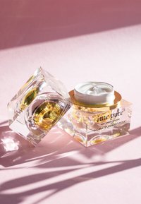 Klarer Glascremebehälter mit einem goldenen Deckel, der weiße Feuchtigkeitscreme enthält, begleitet von einem separaten würfelförmigen Behälter mit gelblichem Inhalt.