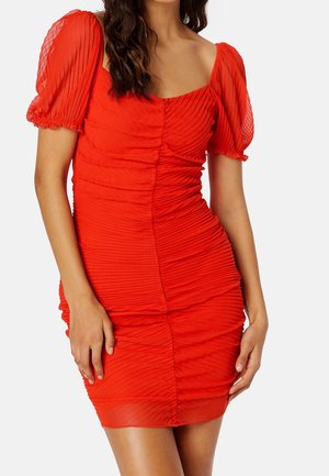 Kobieta ubrana w dopasowaną czerwoną sukienkę z marszczeniami, z krótkimi bufkami, kwadratowym dekoltem i teksturowanym wzorem na białym tle.