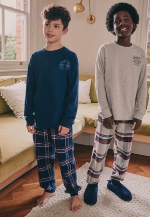 Deux garçons en pantalon de pyjama à carreaux et hauts à manches longues se tiennent dans un salon, l’un pieds nus, l’autre portant des chaussons bleu marine.