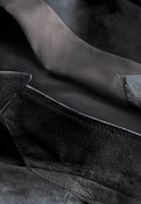 Tela de ante negro con una textura suave, superpuesta sobre una tela tejida más oscura. Los detalles incluyen costuras elaboradas y un acabado suave.