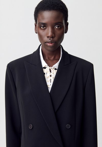 Blazer nero doppiopetto con rever appuntiti, con una camicia bianca con lacci sotto. Tessuto liscio con una silhouette strutturata.