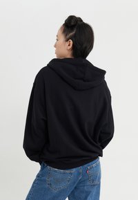 Sort hættetrøje med en løs pasform, der har ribbede manchetter og kant. Kombineret med blå denimjeans, der viser en klassisk fem-lomme stil.