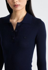 Navy ribbed long-sleeve top with a collared neck, three black buttons, and an embroidered logo on the chest. Durable fabric texture.