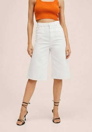 Femme portant un short blanc taille haute jusqu'aux genoux, un débardeur court orange et des sandales noires à talons hauts avec des lanières, debout devant un fond uni.
