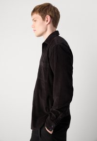 Black corduroy shirt with a button-up design, single chest pocket, and buttoned cuffs, showcasing vertical ribbed texture.