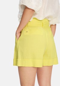 Hochgeschnittene gelbe Shorts mit glatter Textur, seitlichen Taschen und umgeschlagenem Saum am unteren Ende, dezente Hardware-Details auf der Rückseite.