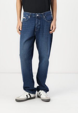 Donkerblauwe denim jeans met een relaxte pasvorm, vijf zakken, zilveren hardware en contrasterende stiksels, gecombineerd met grijs-witte sneakers.
