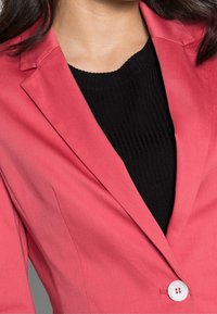 Tætsiddende koralfarvet blazer med skrå kraver, enkelt hvidt knaplukning og en glat tekstur, lagd over en sort ribbet top.