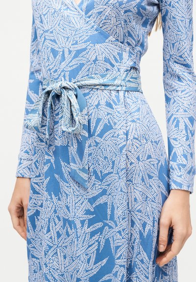 Μπλε φόρεμα με σχέδιο από λευκά πουα φύλλα και ζώνη από ύφασμα δεμένη στη μέση, εμφανίζεται σε ένα άτομο από τους ώμους έως τους γοφούς με τα χέρια χαλαρά στα πλευρά.