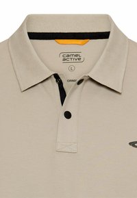 Beige polo-shirt med sorte knapper, sort indvendig krave-snit, og "camel active" logo trykt inde i kraven samt delvist logo på brystet.