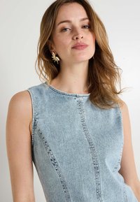Haut sans manches en denim bleu clair avec un col rond et des détails de couture visibles. Modèle portant des boucles d'oreilles en forme de feuille dorée.