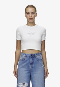 Hvit kortermet t-skjorte med embossert tekst "PECADOR" på forsiden, sammen med lyseblå slitte høyhalsede jeans.