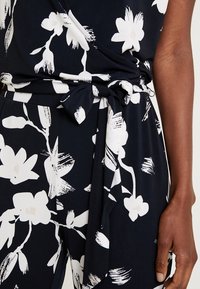 Combinaison florale bleu marine avec motifs de fleurs blanches, un design portefeuille et une ceinture nouée à la taille. Tissu doux avec une texture lisse.