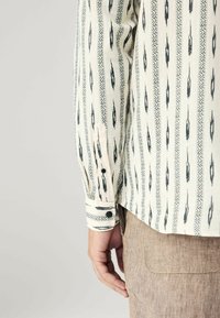 Chemise à manches longues de couleur crème avec des motifs géométriques noirs, dotée d'un poignet à boutons et d'une coupe décontractée. Le tissu semble léger avec une texture douce.