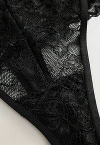 Tanga in pizzo nero con motivi floreali, sezioni trasparenti e bordi in morbido elastico per una finitura delicata.