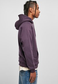 Paarse hoodie met een voorzak, trekkoordcapuchon en geribbelde manchetten. Draagt over een wit shirt, gecombineerd met blauwe spijkerbroeken. Zijaanzicht.