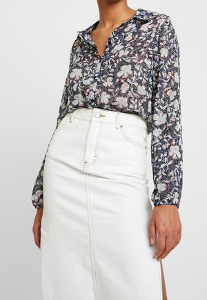 Blusa floral con un fondo oscuro, cuello abotonado y mangas largas. Combinada con una falda de mezclilla blanca con abertura frontal y costuras en contraste.