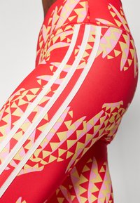 Leggings vermelhos com um padrão geométrico de triângulos rosa e amarelos. Inclui riscas brancas nas laterais. Textura suave.