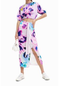 Žena v fialovém a růžovém květinovém crop topu a odpovídající sukni s vysokým rozparkem, držící bílou kabelku a bílé tenisky.