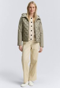 Oliwkowa puchowa kurtka z kapturem, zapinana na zamek błyskawiczny, pod którą znajduje się beżowy sweter na guziki. Do tego kremowe spodnie o szerokich nogawkach i białe buty.