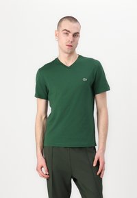 T-shirt verde a maniche corte con scollatura a V realizzato in morbido cotone. Presenta un piccolo logo Lacoste sul lato sinistro del petto. Indossato con pantaloni verdi abbinati.