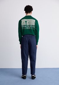 Groene sweatshirt met witte grafische ontwerpen op de achterkant, gecombineerd met marineblauwe broeken. Model draagt zwart-witte sneakers.