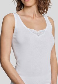 Débardeur blanc en tissu doux, avec une finition en dentelle festonnée ornée de petits motifs floraux et de coutures décoratives à l'encolure.